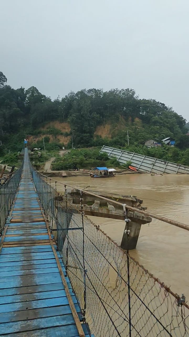 Jembatan gantung berlantai papan biru melintasi Sungai Tamiang, dengan struktur jembatan lain yang tampak rusak di sebelahnya.